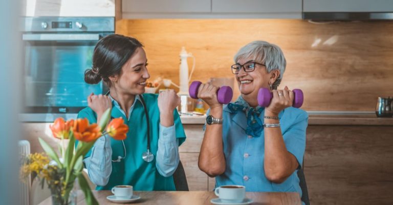 Photo of Nurse helping senior woman in lifting dumbbell at retirement home during the day. Senior woman with dumbbells in rehab with a physiotherapist smiling. Physiotherapist helps elderly woman with healthy dumbbell training in rehab or home in the kitchen while sitting at table.