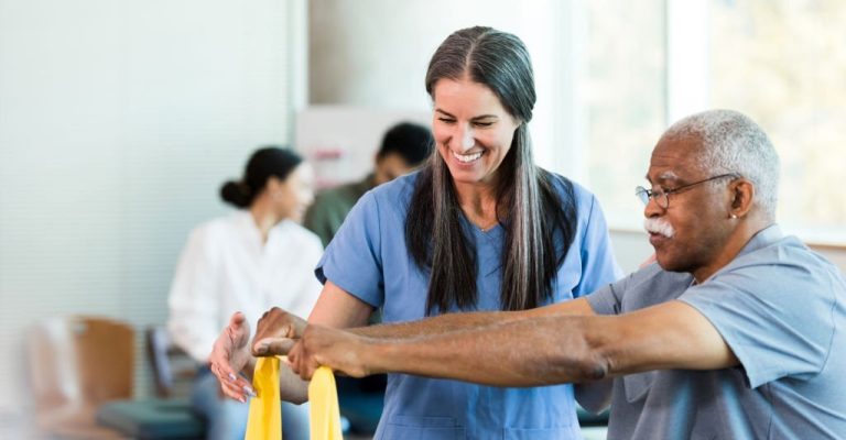 As other therapists work with patients in the background, the mid adult female physical therapist teaches the senior adult man how to use an elastic band to exercise his arms.