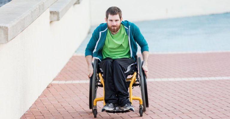 A mid adult man in his 30s in a self-propelled wheelchair, moving toward and looking at the camera, with a serious expression. He is pushing himself uphill, up an inclined walkway. He has cerebral palsy.